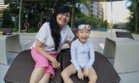 A kid is in swimming costume and goggles is sitting beside her mother by the pool